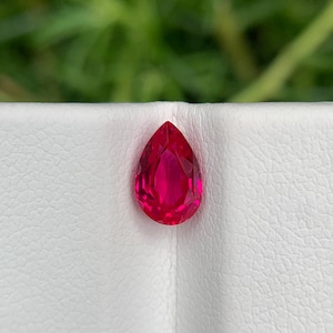 Może przedstawiać: Fasetowany, gruszkowaty, żywy czerwony kamień szlachetny. Kamień jest osadzony na białej powierzchni, z rozmytym zielonym tłem. Szlif kamienia szlachetnego pozwala na odbijanie światła, tworząc efektowny wygląd. Ten kamień szlachetny nadaje się do biżuterii.