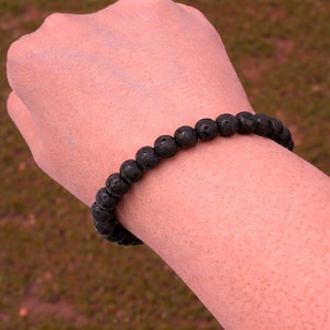 May include: A black lava stone bracelet. The bracelet is made of round, dark gray beads strung together. The bracelet is worn on a wrist, with a blurred background of green and brown.