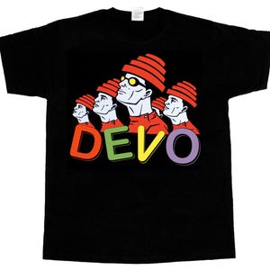 Op de afbeelding: Zwart T-shirt met een pop-art ontwerp van vier figuren met rode hoeden en shirts, en het woord "DEVO" in kleurrijke letters. De figuren kijken omhoog. Het shirt is gemaakt van een zacht materiaal.