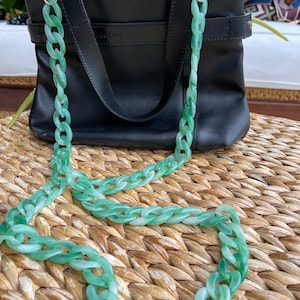 Puede incluir: Una correa de cadena de plástico verde para bolso. La correa está hecha de eslabones gruesos y robustos y está unida a un bolso de cuero negro.