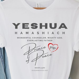 May include: White t-shirt with the text "YESHUA HAMASHIACH" in gray, and "Prince of Peace" in script. A red heart with Hebrew text is below the script. The shirt is hanging on a wooden hanger.