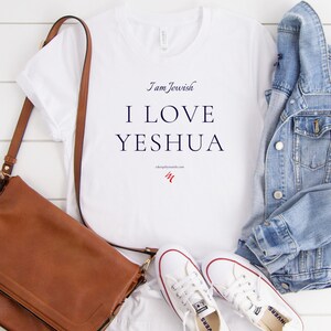 May include: White t-shirt with the text "I am Jewish I LOVE YESHUA". A brown leather shoulder bag, white sneakers, and a light blue denim jacket are also in the image. The items are arranged on a white wooden surface.