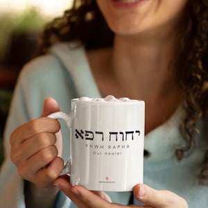 May include: White ceramic mug with the text "YHWH RAPHA Our Healer" in black Hebrew and English lettering. The mug is filled with a white beverage and topped with marshmallows. The mug has a curved handle.