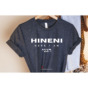 Puede incluir: Camiseta gris oscuro con la palabra "HINENI" en letras mayúsculas blancas grandes, con "HERE I AM" debajo y texto hebreo debajo. La camiseta está colgada en una percha de madera.