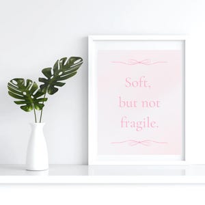 May include: A white framed print with the words "Soft, but not fragile" in pink script. A white vase holds green leaves to the left of the print. The print and vase sit on a white shelf against a white wall.