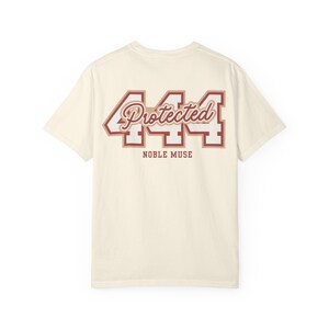 May include: A cream-colored short-sleeve t-shirt featuring a large graphic on the back. The design displays the number 444 in white with a brown outline, overlaid with the word Protected in a brown script font, and the text NOBLE MUSE below.