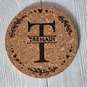 May include: A round cork coaster with a black and white design. The design features a large letter "T" with the name "TREMAIN" written below it. The letter "T" is surrounded by a wreath of leaves and hearts.