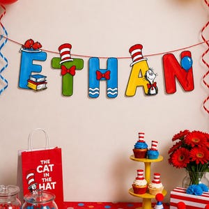 May include: A decorative banner spelling the name ETHAN with colourful letters featuring Cat in the Hat motifs, suspended below red, yellow, and blue balloons. The display includes cupcakes, a red gift bag, and a book titled The Cat in the Hat.