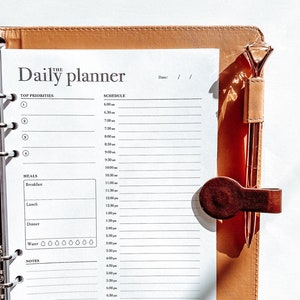 May include: A brown leather daily planner with a white page open to a schedule and meal planning section. The planner has a ring binder and a pen holder with a gold pen.