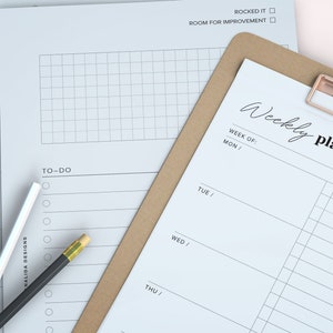 May include: A stack of white paper planner sheets with the words "Weekly plan" and "To-Do" printed on them. A brown clipboard with a rose gold clip holds one sheet. A black pencil and a white pen are on the side.