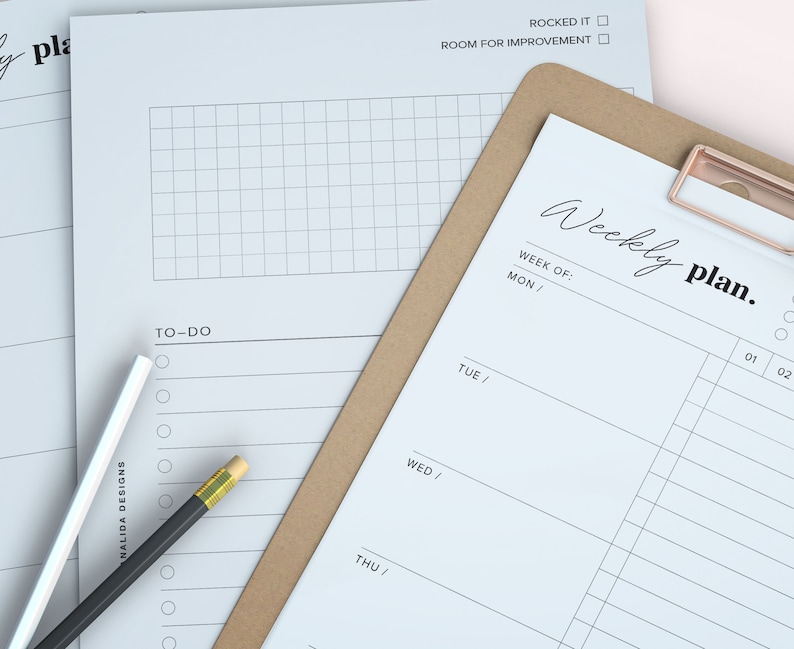 May include: A stack of white paper planner sheets with the words "Weekly plan" and "To-Do" printed on them. A brown clipboard with a rose gold clip holds one sheet. A black pencil and a white pen are on the side.