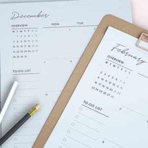 May include: A close-up of a planner with the month of February and December calendars. The planner is on a brown clipboard with a gold clip. A white pencil and a black pencil with a yellow eraser are on the planner.