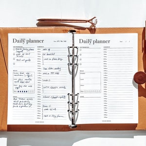 May include: A brown leather bound daily planner with a silver ring binder. The planner has a white page with the text "Daily Planner" and a section for "Top Priorities" and "Schedule". The page is filled with handwritten notes.