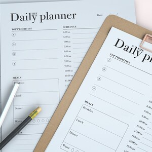 May include: Two white daily planner pages with a brown clipboard on top. The top page has the words "The Daily Planner" and a schedule with times from 6:00 am to 1:00 pm. The bottom page has the words "The Daily Planner" and a schedule with times from 6:00 am to 1:00 pm. Both pages have a section for "Top Priorities" and "Meals".
