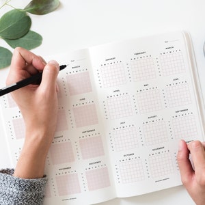 Puede incluir: Una agenda blanca con un dise&ntilde;o de cuadr&iacute;cula para cada mes del a&ntilde;o. La agenda est&aacute; abierta en las p&aacute;ginas de enero a junio. Una persona est&aacute; escribiendo en la secci&oacute;n de marzo con un bol&iacute;grafo negro.