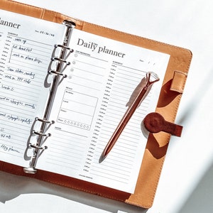May include: A brown leather bound daily planner with a silver ring binder. The planner is open to a page with a schedule for the day, with handwritten notes. A rose gold pen with a diamond-shaped top is resting on the page.