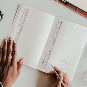 K&ouml;nnte beinhalten: Ein offenes Notizbuch mit einem rosa-wei&szlig;en Gittermuster. Das Notizbuch tr&auml;gt den Titel "Dream Big, Take Action".