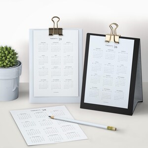 May include: Three white calendars with black text showing the months of the year for 2025 and 2026. The calendars are on a white surface with a small potted plant in the background. The calendars are held together with gold binder clips.