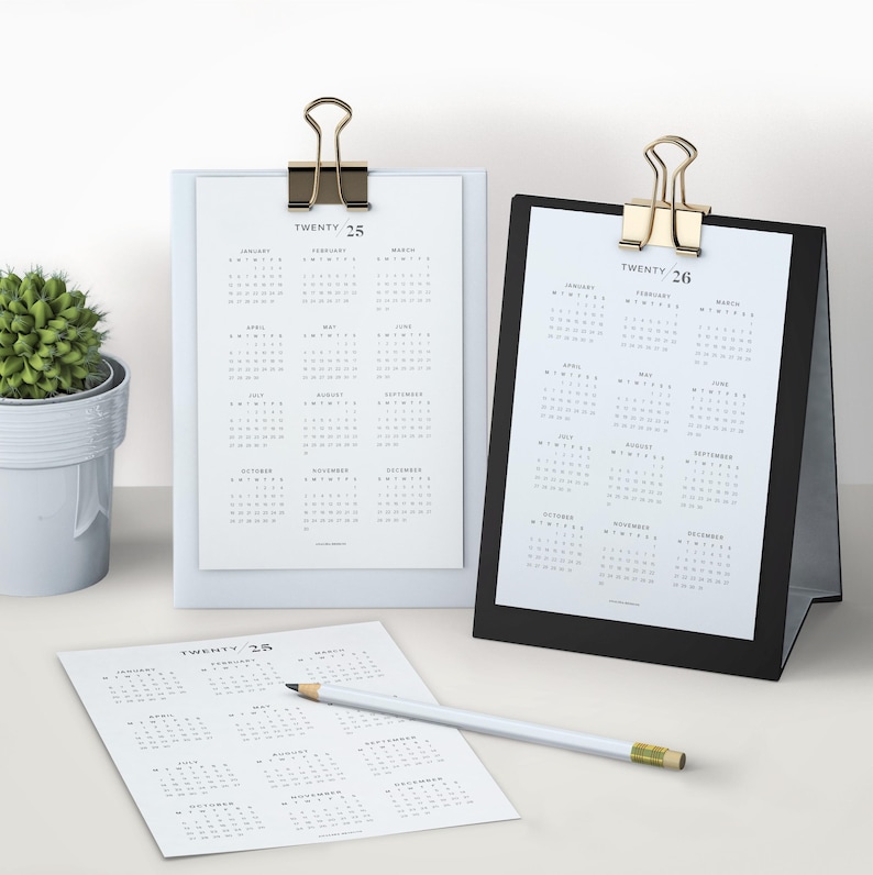 May include: Three white calendars with black text showing the months of the year for 2025 and 2026. The calendars are on a white surface with a small potted plant in the background. The calendars are held together with gold binder clips.
