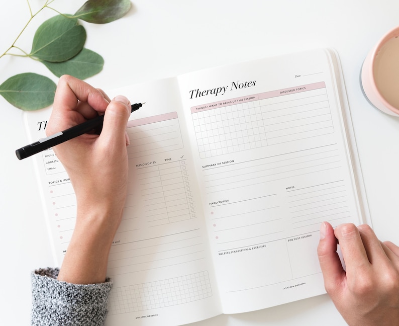 May include: A white and pink therapy notes journal with sections for things to bring up, session dates, time, summary of session, hard topics, notes, helpful suggestions and exercises, and for next session. The journal is open and a person is writing in it with a black pen.