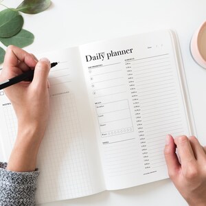 May include: A white daily planner with a black grid pattern and the text "Daily planner" printed on the cover. A person is writing in the planner with a black pen.