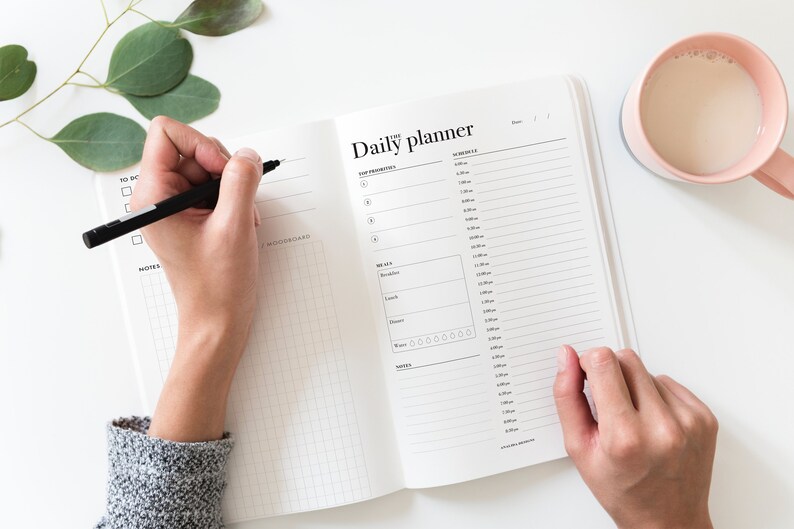 May include: A white daily planner with a black grid pattern and the text "Daily planner" printed on the cover. A person is writing in the planner with a black pen.
