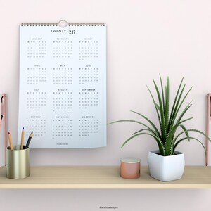 May include: A white calendar with the text "TWENTY 26" printed on the top. The calendar is hanging on a white wall above a wooden shelf. The shelf has a gold metal pencil holder with pencils, a small gold container, and a white planter with a green plant.