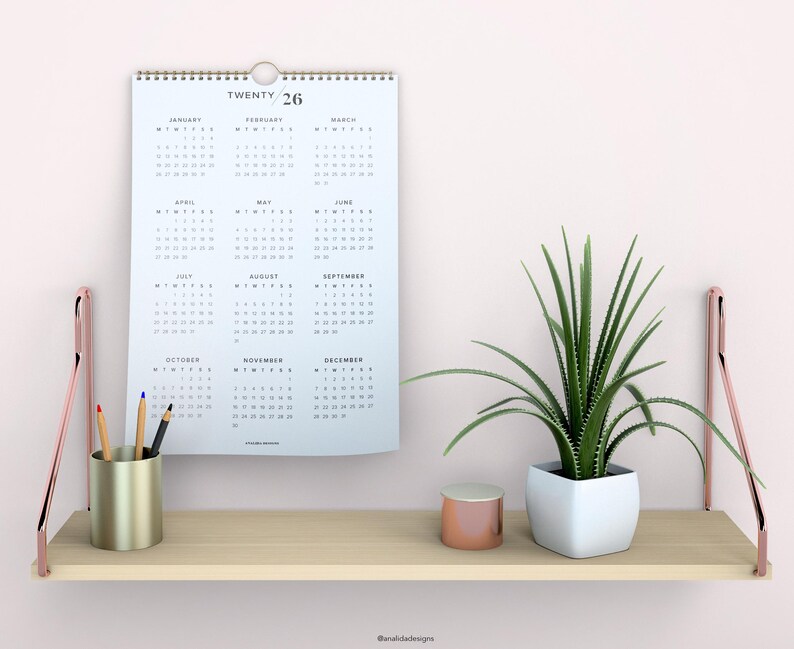 May include: A white calendar with the text "TWENTY 26" printed on the top. The calendar is hanging on a white wall above a wooden shelf. The shelf has a gold metal pencil holder with pencils, a small gold container, and a white planter with a green plant.