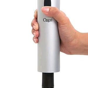 May include: A cylindrical silver electric wine opener with a black top cap and a black oval button on the side. The brand name Ozeri is printed in black text on the front of the metallic body. The device is positioned over the neck of a dark bottle.