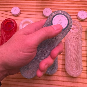 May include: A hand holds a blue, textured, oval-shaped object with a white lid that says "flic". The object is similar to a bottle stopper. There are three other white lids with the word "flic" on them in the background.