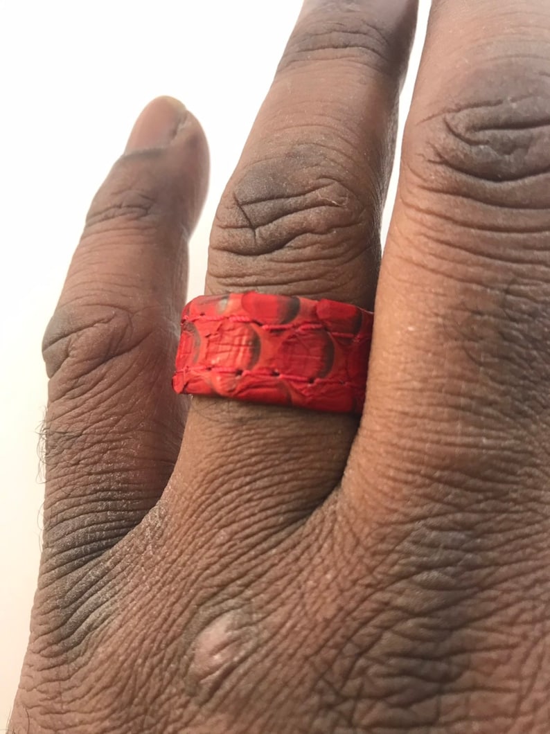 以下が含まれることがあります： 蛇革模様の赤い指輪が指に着用されています。 リングはバンドスタイルで、表面はテクスチャード加工されています。 リングの色は鮮やかな赤色で、濃い茶色のアクセントが付いています。