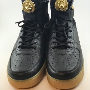 May include: Pair of black high-top sneakers with gold lion head accents on the tongue. The sneakers have black laces, a perforated toe box, and a tan sole. The shoes are made of leather and fabric.