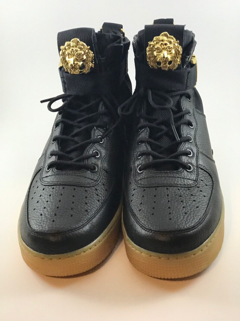 May include: Pair of black high-top sneakers with gold lion head accents on the tongue. The sneakers have black laces, a perforated toe box, and a tan sole. The shoes are made of leather and fabric.