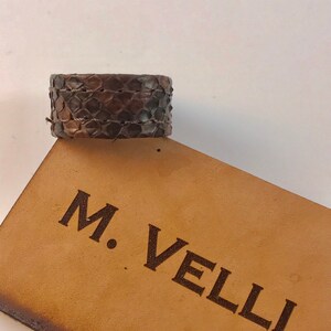 以下が含まれることがあります： ヘビ革模様の幅広の茶色の革製カフブレスレット。ブレスレットは細い織りコードで固定されています。「M. Velli」の文字が薄茶色の革タグに印刷されています。