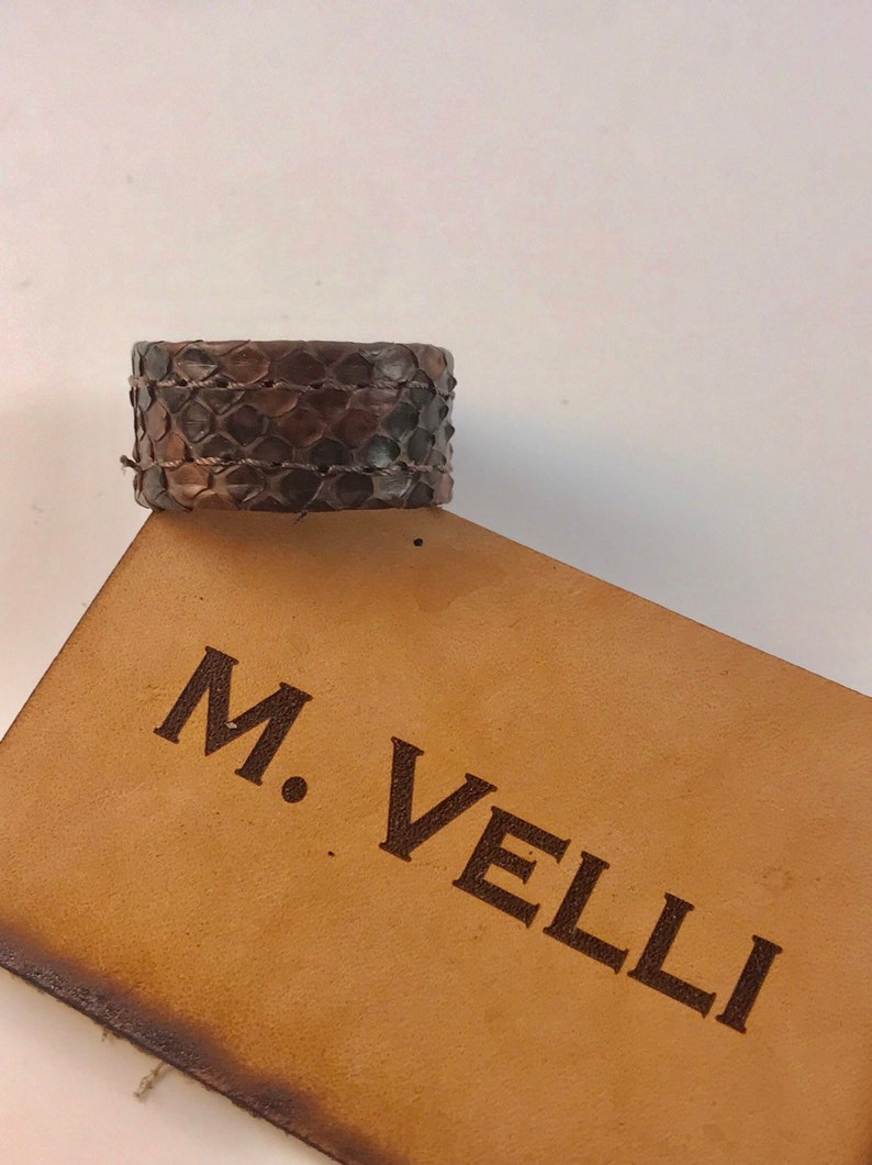 以下が含まれることがあります： ヘビ革模様の幅広の茶色の革製カフブレスレット。ブレスレットは細い織りコードで固定されています。「M. Velli」の文字が薄茶色の革タグに印刷されています。
