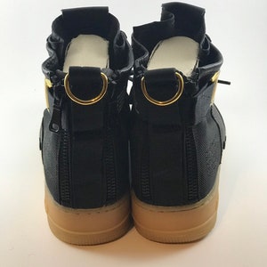 May include: Pair of black high-top sneakers with tan soles. Features include gold-colored metal accents, zippers, and a unique design. The sneakers have a modern, urban aesthetic.