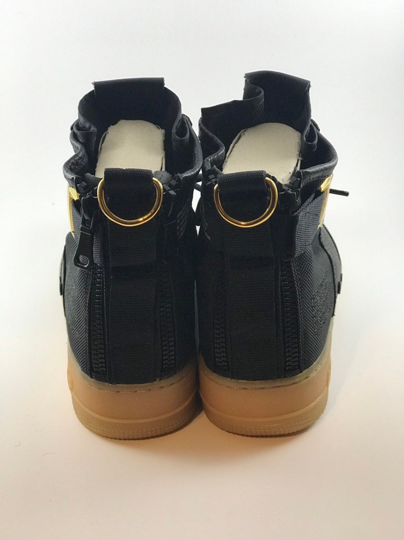 May include: Pair of black high-top sneakers with tan soles. Features include gold-colored metal accents, zippers, and a unique design. The sneakers have a modern, urban aesthetic.