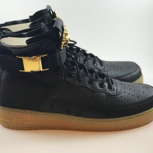 May include: Black high-top sneakers with a textured leather upper and a tan rubber sole. The sneakers feature gold-colored accents on the strap and heel, and a lace-up closure. The word "AIR" is visible on the sole.