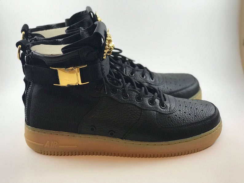 May include: Black high-top sneakers with a textured leather upper and a tan rubber sole. The sneakers feature gold-colored accents on the strap and heel, and a lace-up closure. The word "AIR" is visible on the sole.