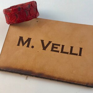 以下が含まれることがあります： 赤い革のブレスレットは、鱗模様で、薄茶色の革の長方形の上に置かれています。長方形には、濃い茶色の文字で「M. VELLI」と書かれています。革の端はわずかに暗くなっています。