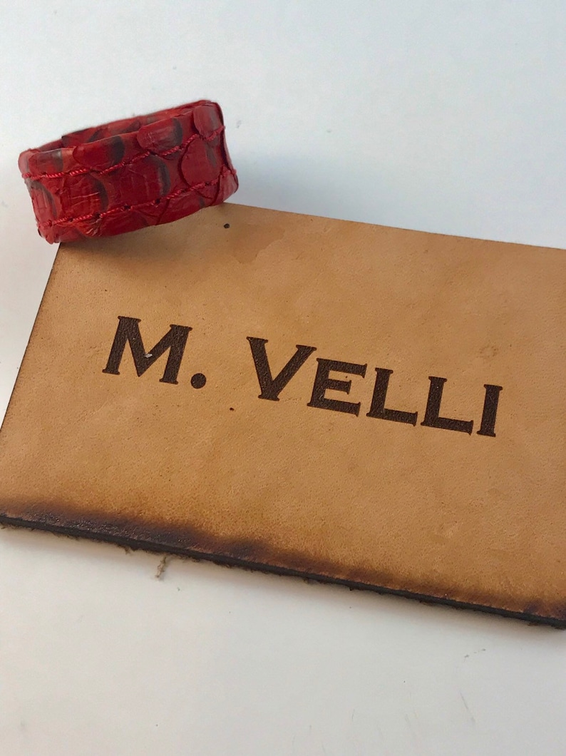 以下が含まれることがあります： 赤い革のブレスレットは、鱗模様で、薄茶色の革の長方形の上に置かれています。長方形には、濃い茶色の文字で「M. VELLI」と書かれています。革の端はわずかに暗くなっています。