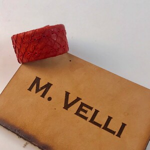 以下が含まれることがあります： 織り模様の赤い革製ブレスレットが、タン色の革製タグの上に置かれています。タグには、濃い茶色のフォントで「M. VELLI」の文字が刻印されています。ブレスレットは鮮やかな赤色で、小さくテクスチャ加工されたパーツでできているようです。