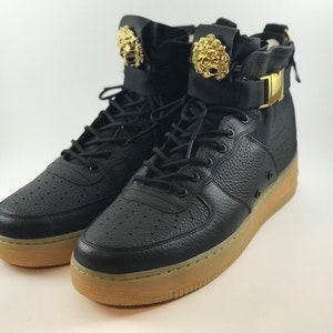 May include: Pair of black high-top sneakers with gold lion head accents on the ankle straps. The sneakers have black laces, a perforated leather upper, and a tan rubber sole. A gold buckle secures the ankle strap.