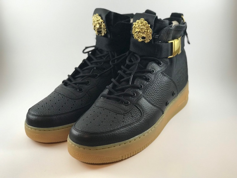 May include: Pair of black high-top sneakers with gold lion head accents on the ankle straps. The sneakers have black laces, a perforated leather upper, and a tan rubber sole. A gold buckle secures the ankle strap.