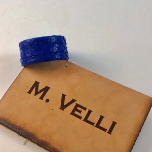 以下が含まれることがあります： 鮮やかな青色の革製ブレスレット。鱗のような模様のテクスチャが特徴です。ブレスレットは、「M. VELLI」の文字が刻まれた、明るい茶色の革の表面に置かれています。