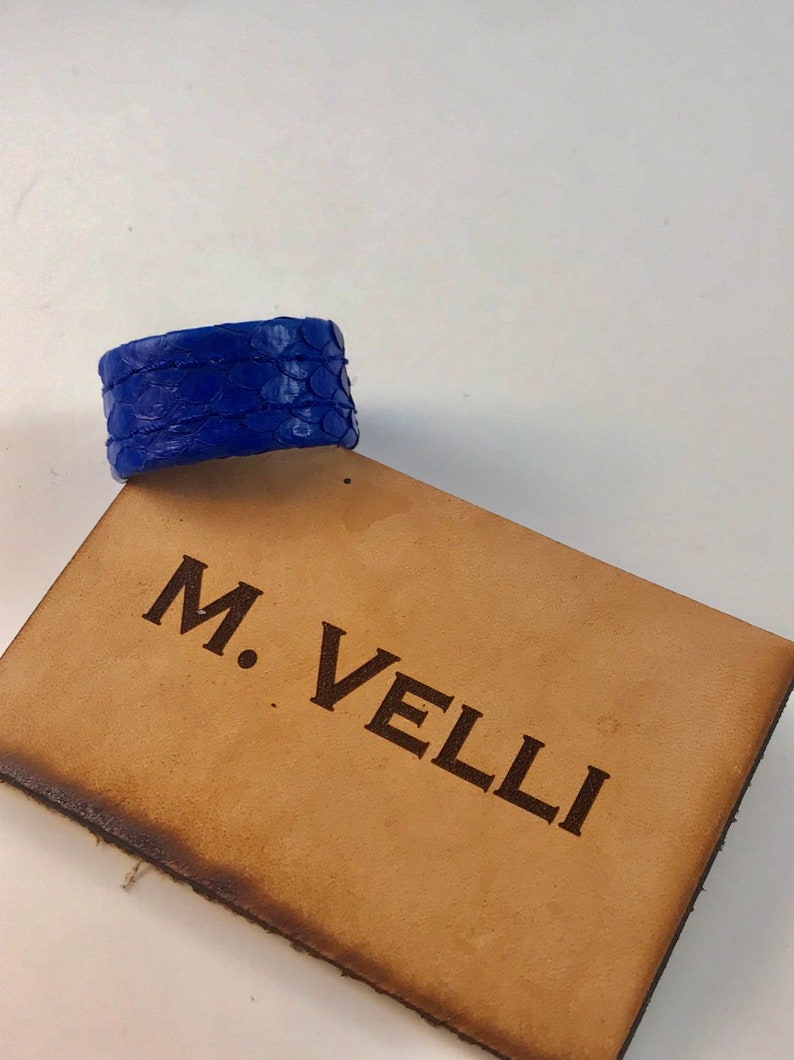 以下が含まれることがあります： 鮮やかな青色の革製ブレスレット。鱗のような模様のテクスチャが特徴です。ブレスレットは、「M. VELLI」の文字が刻まれた、明るい茶色の革の表面に置かれています。