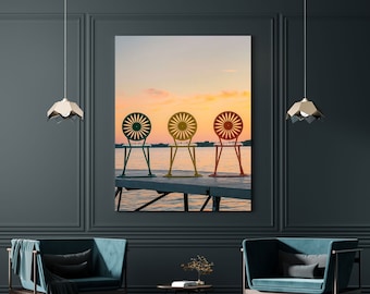 May include: Three colorful chairs with a circular back design sit on a wooden dock overlooking a lake at sunset. The chairs are green, yellow, and red.