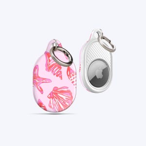Op de afbeelding: Twee AirTag-houders. Eén is roze met een schelp- en zeesterpatroon, de andere is wit met een zilveren ring en een zwart Apple-logo. Beide hebben zilveren sleutelringen.