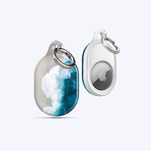 Könnte beinhalten: Zwei AirTag-Halter. Einer zeigt eine Strandszene mit weißen Wellen und blauem Wasser. Der andere ist weiß mit einem türkisfarbenen Rand und einem silbernen Ring. Beide haben silberne Schlüsselringe.