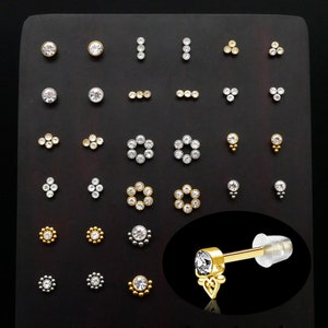 Op de afbeelding: Een display met verschillende gouden en zilveren lichaamsieraden met heldere kristallen accenten. De sieraden omvatten studs, staven en ringen met verschillende ontwerpen. De display is op een zwarte achtergrond.